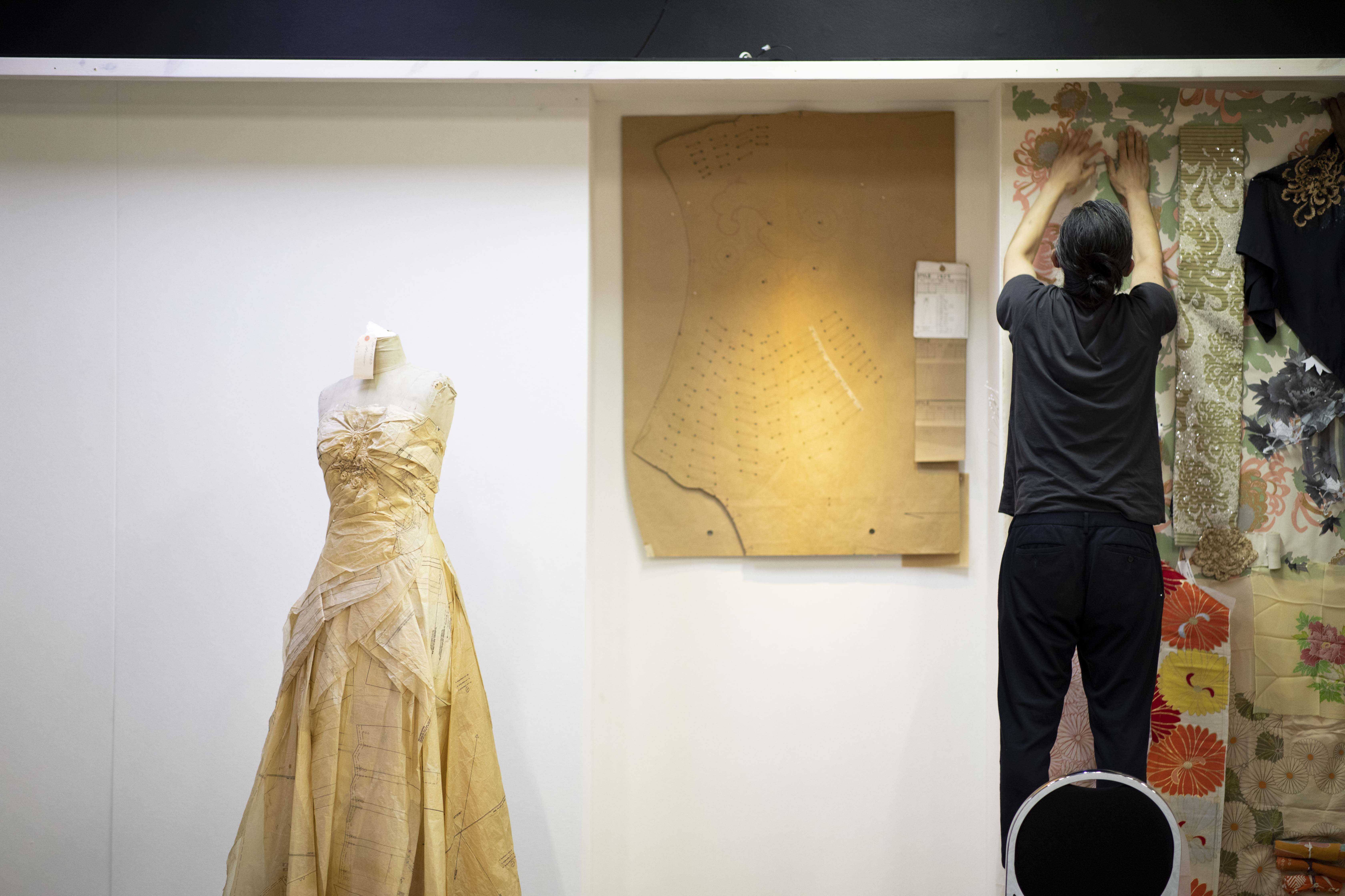 A man standing on a chair pinning fabric and other objects on a large board. To his left is a brown cardboard pattern piece and a prototype evening dress made from paper tissue pinned to a dress form.